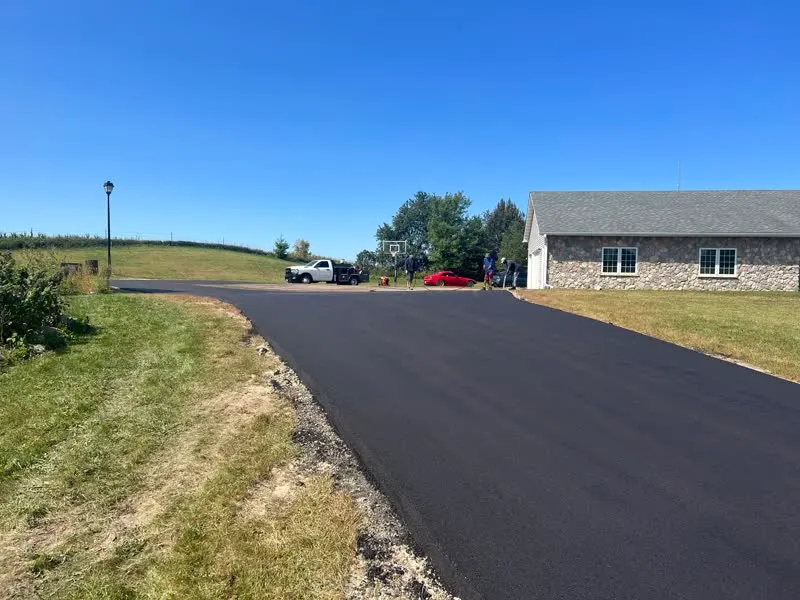 Freshly paved black asphalt driveway leading to a stone house with green lawns under a clear blue sky